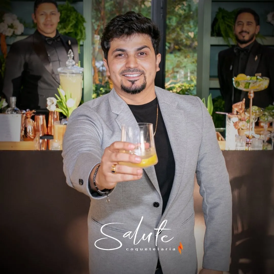 Retrato de Cleiton Venditi erguendo um drink, com a equipe do bar ao fundo